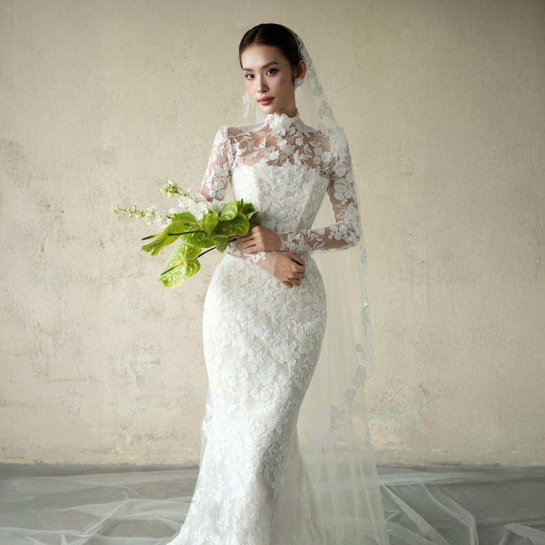 Puede incluir: Vestido de novia de encaje blanco con mangas largas y cuello alto. El vestido tiene un corpi&ntilde;o ajustado y una falda de sirena. Un velo transparente y un ramo de flores blancas y verdes completan el look nupcial.