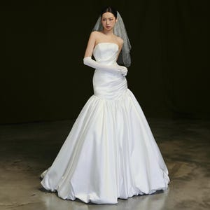 Può includere: Un abito da sposa in raso bianco senza spalline, con silhouette a sirena. L'abito presenta un corpetto aderente, una vita arricciata e una gonna svasata. La sposa indossa lunghi guanti bianchi e un velo.