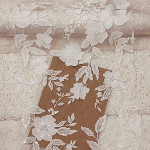 Puede incluir: Tela de encaje floral blanco con flores y hojas bordadas, adornada con lentejuelas. Este delicado encaje es ideal para ropa de novia, vestidos o proyectos de manualidades. El encaje est&aacute; superpuesto a una superficie marr&oacute;n.