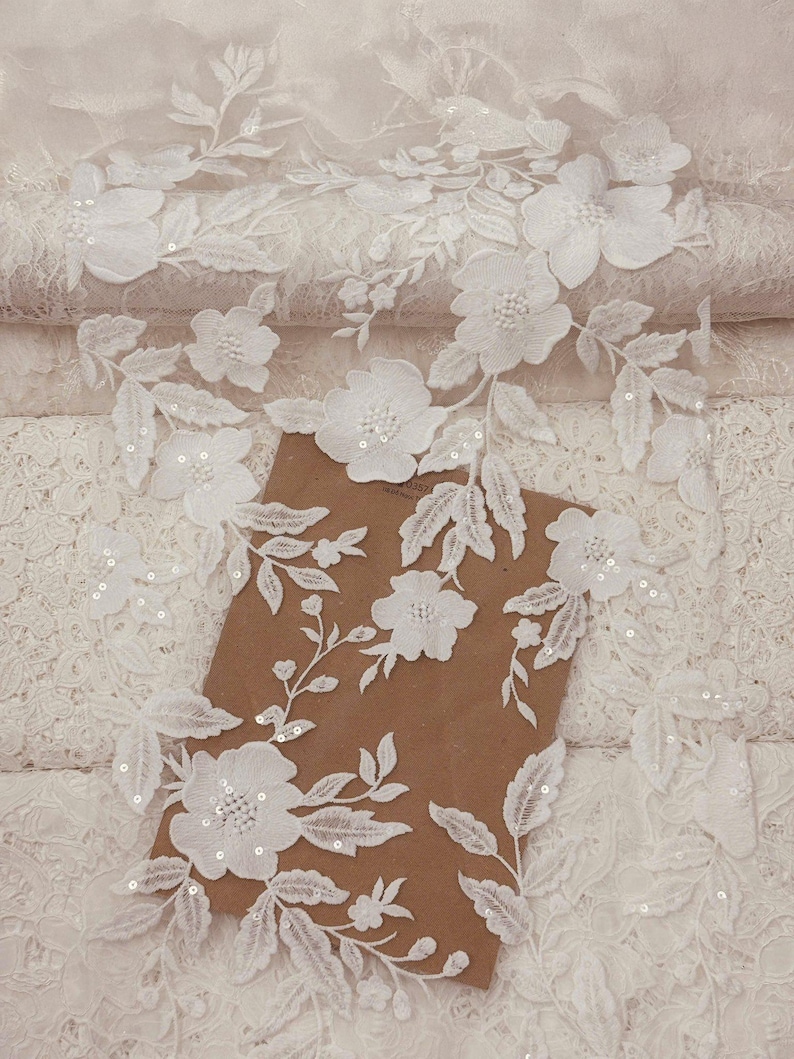 Puede incluir: Tela de encaje floral blanco con flores y hojas bordadas, adornada con lentejuelas. Este delicado encaje es ideal para ropa de novia, vestidos o proyectos de manualidades. El encaje est&aacute; superpuesto a una superficie marr&oacute;n.