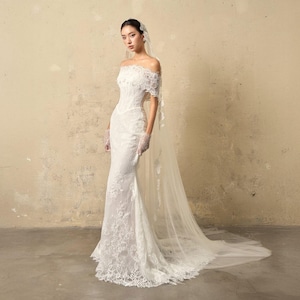 May include: A white, off-the-shoulder wedding dress with intricate lace detailing. The gown features a fitted bodice, a mermaid skirt, and a long, flowing train. The bride is wearing a veil and lace gloves.