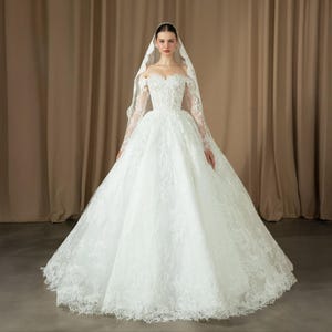 Può includere: Un abito da sposa bianco con scollatura a spalle scoperte e maniche lunghe. L'abito presenta intricati dettagli in pizzo sul corpetto e sulla gonna, con una silhouette ampia e fluida. Un velo abbinato drappeggia dalla testa, completando l'ensemble nuziale.