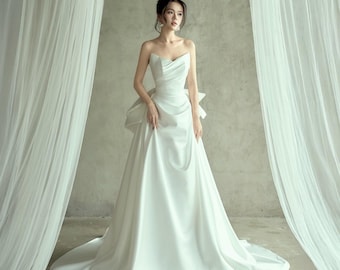 May include: A strapless white satin wedding gown with a flowing A-line skirt and a long train. The bodice features a sweetheart neckline and a bow detail at the back. The dress is displayed in a studio setting with white curtains.