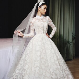 Può includere: Un abito da sposa in pizzo bianco con maniche lunghe e collo alto. L'abito presenta una gonna ampia e una fila di bottoni sul davanti. Un velo trasparente e un copricapo floreale completano il look da sposa.