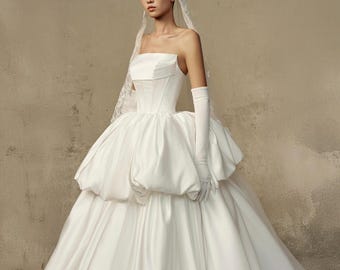 May include: A strapless white wedding gown with a structured bodice and a voluminous skirt. The dress features a unique bubble-like design around the waist and is paired with long white gloves and a delicate veil. The gown is made of a smooth, satin-like fabric.