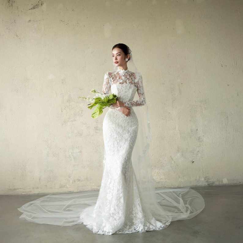 Puede incluir: Un vestido de novia de encaje blanco con mangas largas y cuello alto. El vestido tiene un corpi&ntilde;o ajustado y una falda de sirena. Un velo largo y transparente se extiende por detr&aacute;s del vestido. La persona sostiene un ramo de flores blancas y verdes.