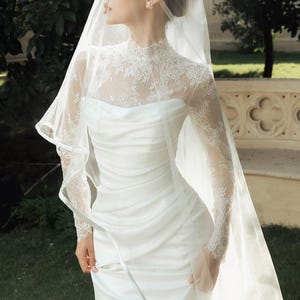 Peut inclure: Une robe blanche longue jusqu'au sol avec un corsage fronc&eacute; et un empi&egrave;cement en dentelle &agrave; col montant avec des manches longues. Le v&ecirc;tement est associ&eacute; &agrave; un voile en tulle transparent qui tombe sur la t&ecirc;te et les &eacute;paules en couches.