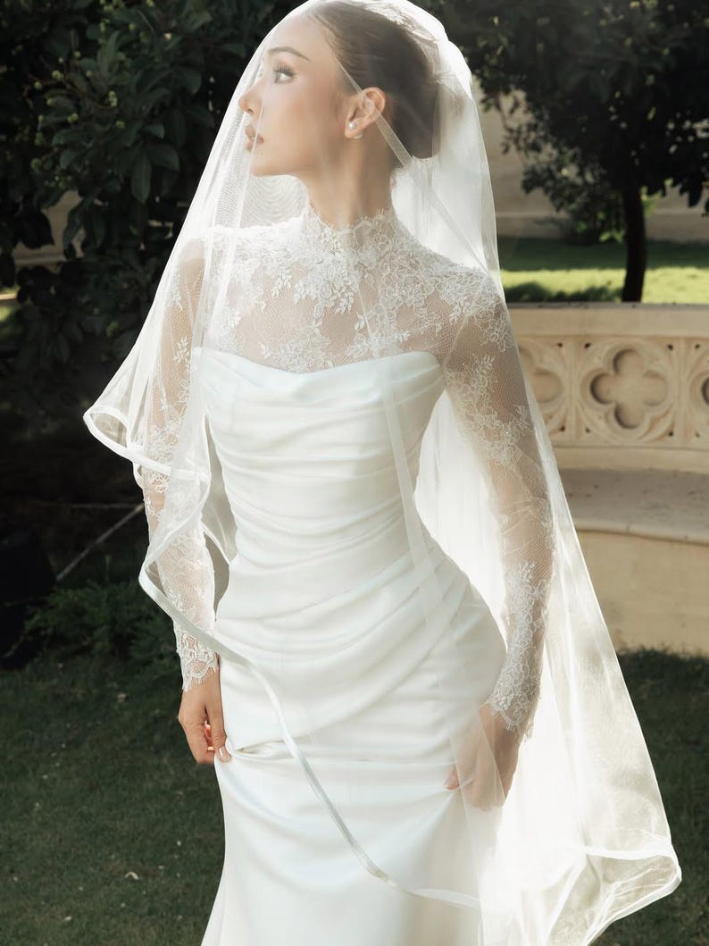 Peut inclure: Une robe blanche longue jusqu'au sol avec un corsage fronc&eacute; et un empi&egrave;cement en dentelle &agrave; col montant avec des manches longues. Le v&ecirc;tement est associ&eacute; &agrave; un voile en tulle transparent qui tombe sur la t&ecirc;te et les &eacute;paules en couches.