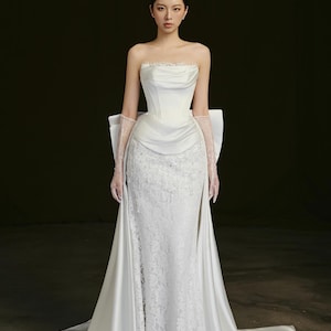 Può includere: Un abito da sposa bianco senza spalline con corpetto aderente e gonna fluida. L'abito presenta un rivestimento in pizzo sulla gonna e un grande fiocco sul retro. La modella indossa guanti di pizzo trasparenti.