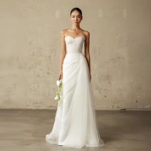 Può includere: Un abito da sposa bianco senza spalline con corpetto aderente e gonna fluida. L'abito presenta uno scollo a cuore ed è abbinato a un piccolo bouquet di fiori bianchi. La modella è in piedi davanti a un muro dai toni neutri.
