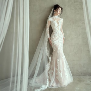 May include: A white, long-sleeved wedding dress with intricate floral lace detailing. The dress features a high neckline, a fitted bodice, and a flowing mermaid skirt. A sheer veil with lace trim cascades down the back. The dress is a classic bridal gown.