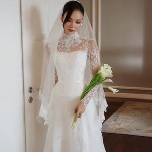 Può includere: Abito da sposa bianco con scollatura alta in pizzo e maniche lunghe in pizzo. L'abito presenta un corpetto aderente e una gonna fluida. Un velo trasparente e un bouquet di calle bianche completano il look da sposa.