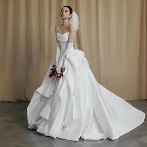 Può includere: Un abito da sposa bianco senza spalline con gonna voluminosa e lungo strascico. La sposa indossa lunghi guanti bianchi e tiene in mano un piccolo bouquet di fiori rossi. Un velo corto è attaccato alla parte posteriore della testa.