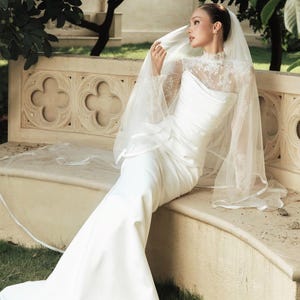 Peut inclure: Une robe de mari&eacute;e blanche longue jusqu'au sol avec un corsage en dentelle &agrave; col montant et des manches longues, associ&eacute;e &agrave; une jupe unie et lisse. Un voile l&eacute;ger et transparent &agrave; bordure finie recouvre les &eacute;paules et la t&ecirc;te de la tenue.