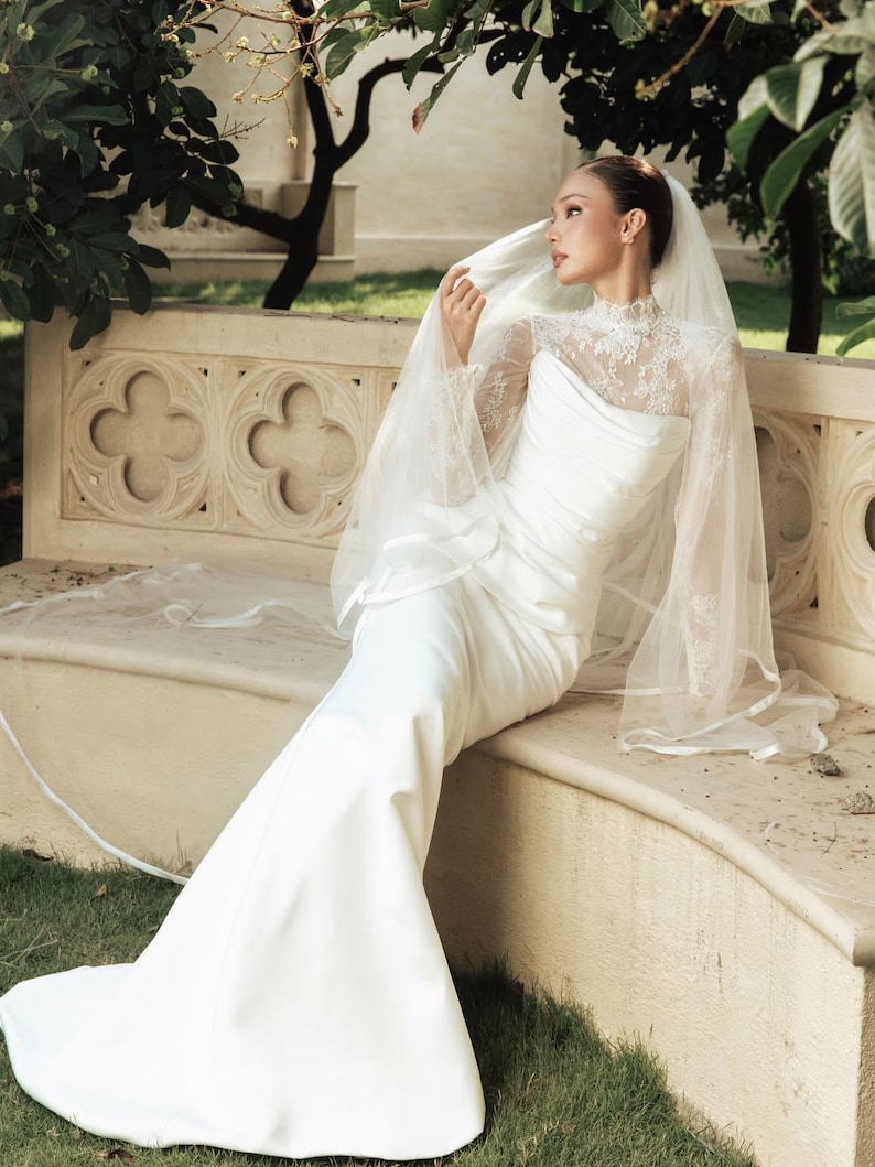 Peut inclure: Une robe de mari&eacute;e blanche longue jusqu'au sol avec un corsage en dentelle &agrave; col montant et des manches longues, associ&eacute;e &agrave; une jupe unie et lisse. Un voile l&eacute;ger et transparent &agrave; bordure finie recouvre les &eacute;paules et la t&ecirc;te de la tenue.