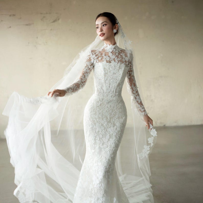 Puede incluir: Vestido de novia blanco de manga larga y cuello alto. El vestido presenta una silueta de sirena y est&aacute; adornado con encaje floral. Un velo transparente con detalles florales est&aacute; adjunto.