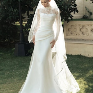 Peut inclure: Une robe de mari&eacute;e blanche longue jusqu'au sol avec un corsage en dentelle &agrave; col montant, des manches longues et une jupe en satin drap&eacute;. La robe est accompagn&eacute;e d'un long voile transparent qui tombe sur les &eacute;paules et jusqu'au sol.