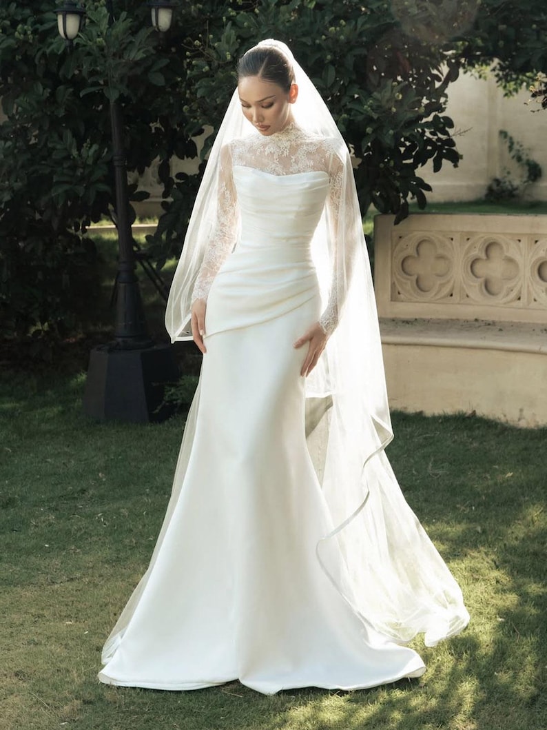 Peut inclure: Une robe de mari&eacute;e blanche longue jusqu'au sol avec un corsage en dentelle &agrave; col montant, des manches longues et une jupe en satin drap&eacute;. La robe est accompagn&eacute;e d'un long voile transparent qui tombe sur les &eacute;paules et jusqu'au sol.