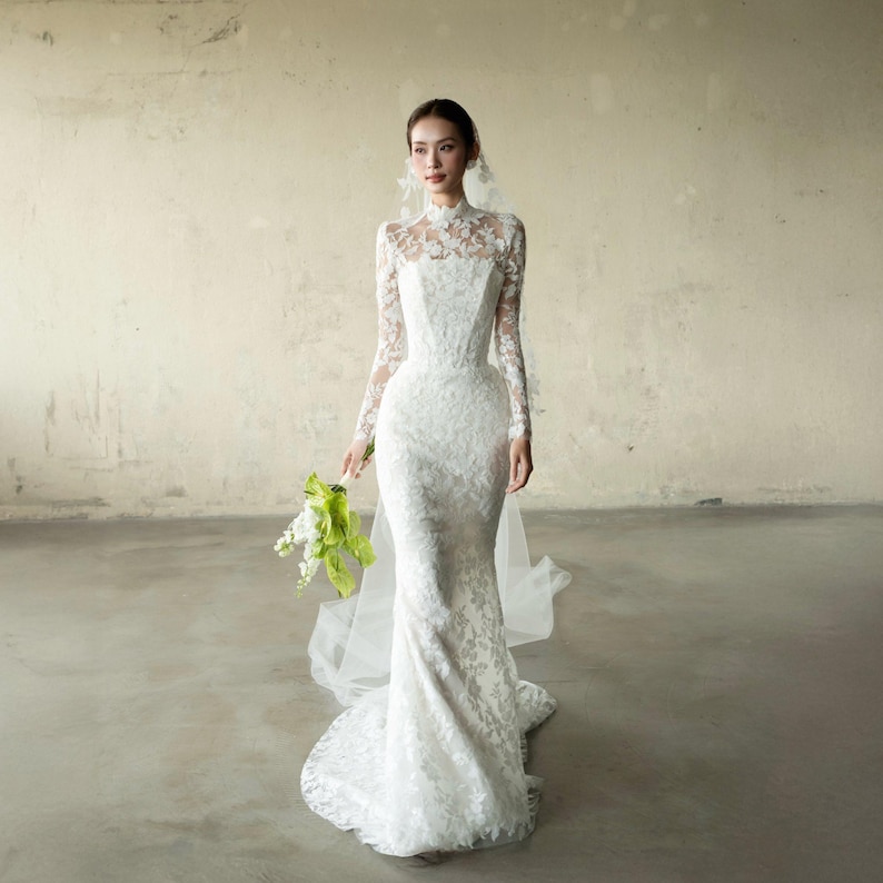 Puede incluir: Un vestido de novia blanco, largo hasta el suelo, con cuello alto y mangas largas. El vestido presenta intrincados detalles de encaje y una silueta ajustada. Un velo transparente y un ramo de flores blancas y verdes completan el look nupcial.