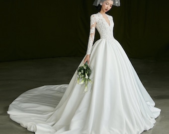 May include: A white wedding dress with a long train. The dress features a V-neckline, long lace sleeves, and a full skirt. A white hat with a veil is worn. The bride holds a bouquet of white flowers and greenery.