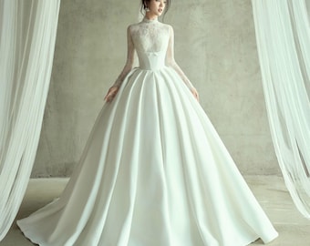 May include: A white wedding gown with a high neckline and long sleeves. The bodice features lace detailing and a small bow. The skirt is full and flowing, creating a classic and elegant silhouette. The dress is made of a smooth, satin-like fabric.