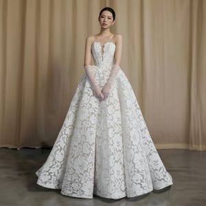 Può includere: Un abito da sposa bianco senza spalline con scollatura a cuore e gonna ampia. L'abito presenta intricati dettagli in pizzo floreale ed è abbinato a guanti lunghi e trasparenti. L'abito lungo crea una silhouette classica ed elegante.