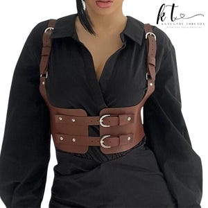 May include: Brown leather harness with silver buckles and rivets. The harness has shoulder straps and a waist belt with two buckles. The text "kt Knaughty Threads Fearless Fashionistas" is in the upper right corner.