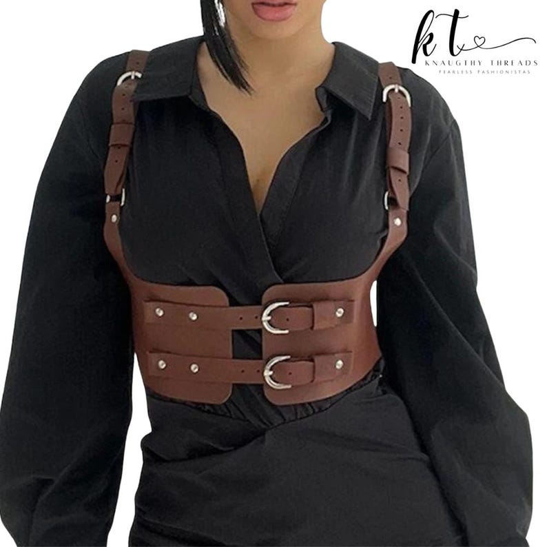 May include: Brown leather harness with silver buckles and rivets. The harness has shoulder straps and a waist belt with two buckles. The text "kt Knaughty Threads Fearless Fashionistas" is in the upper right corner.