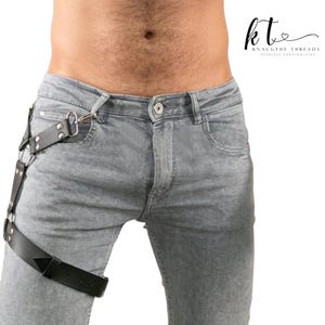 May include: Black leather thigh harness with silver hardware attached to grey denim jeans. The harness has multiple straps and a geometric design at the thigh. The jeans have a button closure and classic five-pocket styling.