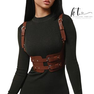 May include: Brown leather harness with silver buckles and straps. The harness has a chest plate and two waist straps. The text "kt" and "KNAUGHTY THREADS FEARLESS FASHIONISTAS" is visible in the upper right corner.