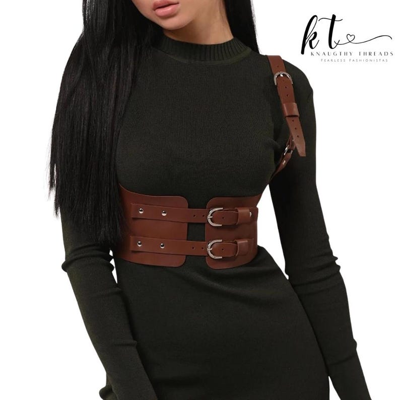 May include: Brown leather harness belt with two silver buckles and a shoulder strap. The belt is designed to cinch the waist and features silver rivets. The image includes the text "kt KNAUGHTY THREADS FEARLESS FASHIONISTAS".