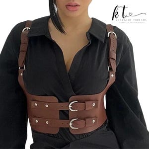 May include: Brown leather harness with silver-tone hardware. The harness features adjustable straps and two buckle closures on the waist. The design includes decorative rivets. The brand logo "kt KNAUGHTY THREADS FEARLESS FASHIONISTAS" is visible in the upper right corner.