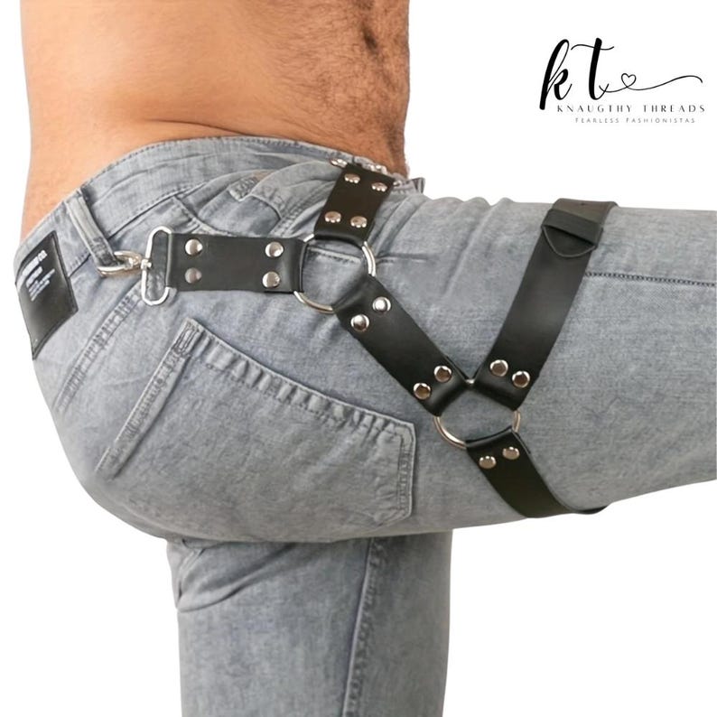 May include: Black leather harness with silver metal rings and rivets. The harness has a Y-shaped design and is attached to a belt loop with a silver clasp. The brand name "KNAUGHTY THREADS" is visible in the upper right corner.