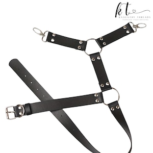 May include: Black leather harness with silver metal hardware, including rings, rivets, and clasps. The harness features adjustable straps and a buckle closure. The brand name "Knaughty Threads" is visible in the upper right corner.
