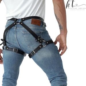 May include: Black leather harness with silver-tone metal rings and buckles. The harness is worn over blue jeans and features straps that connect around the waist and thighs. The brand name "KNAUGHTY THREADS" is visible in the upper right corner.