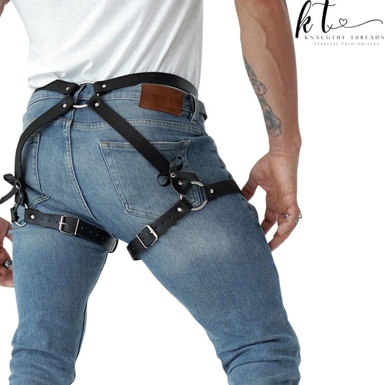 May include: Black leather harness with silver-tone metal rings and buckles. The harness is worn over blue jeans and features straps that connect around the waist and thighs. The brand name "KNAUGHTY THREADS" is visible in the upper right corner.