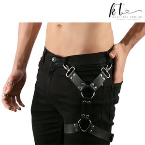 May include: Black leather harness with silver metal hardware. The harness has a Y-shaped design with adjustable straps and a large ring at the centre. The image shows the harness worn over black trousers.