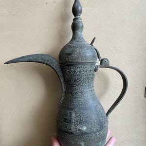 Könnte beinhalten: Eine hohe Kaffeekanne aus dunklem Metall mit einem langen, geschwungenen Ausguss, einem Klappdeckel mit spitzem Aufsatz und einem gebogenen Griff. Die Oberfläche ist mit detaillierten floralen Gravuren und arabischer Kalligrafie verziert.