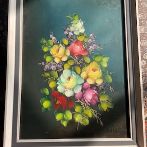 May include: Oil painting of a floral bouquet in a silver frame. The bouquet features yellow, red, pink, and white roses with green leaves and purple accents against a dark teal background. The artist's signature, "Lucas", is visible.