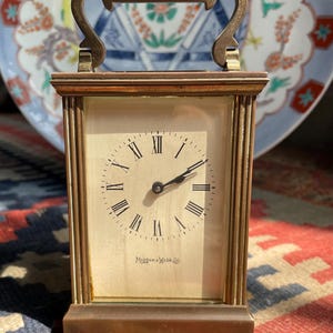 May include: A rectangular brass-framed clock with a cream-colored face and black Roman numerals. The clock has a handle and the brand name "Mappin & Webb Ltd" is printed on the face. The clock is set on a patterned rug.