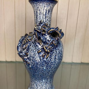 Puede incluir: Un jarrón de cerámica azul y blanco con un diseño de dragón. El jarrón tiene una superficie texturizada y una parte superior acampanada. El dragón está esculpido en relieve y tiene detalles dorados. El jarrón mide aproximadamente 25 cm de alto.