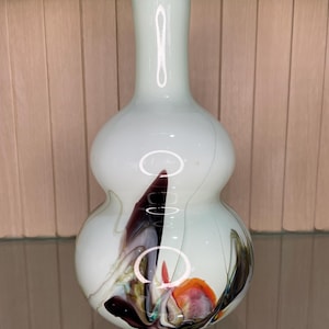 Könnte beinhalten: Eine hellgrüne Glasvase mit bauchigem Körper und schlankem Hals. Die Vase weist abstrakte, farbenfrohe Wirbel aus Rot, Schwarz und Orange auf. Die Vase wird auf einem Glasregal ausgestellt.