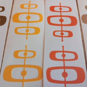 Op de afbeelding: Close-up van verschillende stickers in mid-century modern stijl in tinten bruin, oranje en geel. Elke sticker heeft een verticaal ontwerp met verbonden ovale en cirkelvormige vormen. De stickers staan op een witte achtergrond.