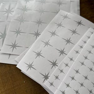 Może przedstawiać: Kilka arkuszy białego papieru z powtarzającym się wzorem szarych gwiazd. Gwiazdy różnią się rozmiarem i są równomiernie rozmieszczone. Obraz przedstawia kilka arkuszy ułożonych razem.