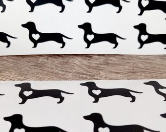 May include: Sheet of black dachshund dog stickers with a white heart-shaped cutout on the dog's body. The stickers are arranged in rows on a white background. The background is a light brown wood grain.