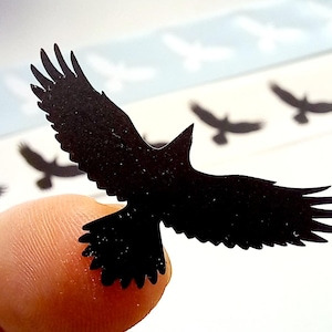 May include: A small, black, die-cut sticker in the shape of a bird in flight with spread wings. The sticker features a solid black silhouette of a bird, and it is part of a larger sheet containing multiple identical bird stickers in a row.