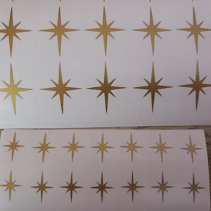 Puede incluir: Hojas de pegatinas de estrellas doradas sobre papel blanco. Cada estrella tiene ocho puntas y un acabado metálico. Las pegatinas están dispuestas en filas y columnas, para manualidades o decoraciones.