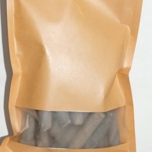 Pode incluir: Uma sacola de papel marrom com uma janela transparente, selada com um z&iacute;per. A janela revela uma cole&ccedil;&atilde;o de objetos cil&iacute;ndricos cinzentos. A sacola &eacute; feita de papel marrom claro.
