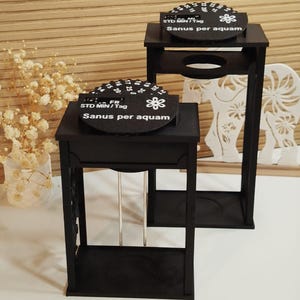 Könnte beinhalten: Zwei schwarze Holzständer mit einer kreisförmigen Oberseite mit dem Text "STD MIN/Tag" und "Sanus per aquam". Die Ständer haben eine rechteckige Basis und ein dekoratives Blumendesign. Die Ständer stehen auf einer weißen Oberfläche.