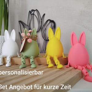 Könnte beinhalten: Ein Set aus vier bunten Osterhasen-Figuren. Die Hasen sind eiförmig mit beweglichen Beinen und Ohren, in Weiß, Grün, Gelb und Pink. Der Text "personalisierbar" und "Set Angebot für kurze Zeit" ist sichtbar.